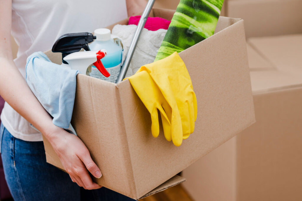 Person holding a cardboard box filled with cleaning supplies and yellow gloves for move-in or move-out cleaning.